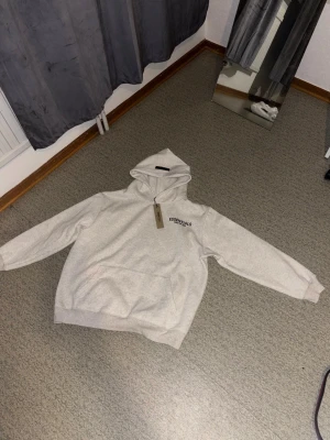 Grå Essentials Fear of God hoodie - Säljer en ljusgrå hoodie från Essentials Fear of God med svart logga på bröstet och stor text på ryggen. Hoodien har en klassisk känguruficka och huva. Tillverkad i mjukt bomullsmaterial som är perfekt för en avslappnad streetwear-look.