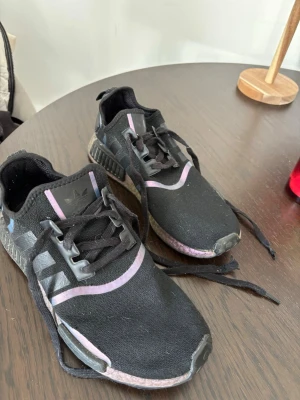 Svarta Adidas NMD sneakers - Säljer ett par svarta Adidas NMD sneakers med klassiska tre ränder på sidan och lila detaljer runt sulan och ovandelen. Skorna har en stickad mesh-överdel och svarta snören. Perfekta för dig som gillar streetwear och bekväma skor.
