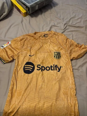 Barcelona bortatröja guld Nike - Snygg FC Barcelona bortatröja i guld med subtilt kartmönster, svart Nike-logga och klubbmärke på bröstet. Spotify-logga framtill och La Liga-märke på ärmen. Tillverkad i lätt och ventilerande material, perfekt för fotboll eller streetwear.