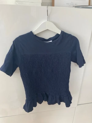 Mörkblå topp med volang från Gina Tricot - Supersöt mörkblå topp från Gina Tricot med volangkant nedtill. Kortärmad modell i mjuk bomull, perfekt nu till sommren.