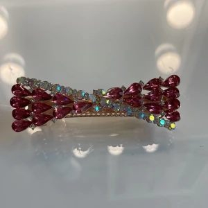 Rosa glittrig hårklämma med stenar - Superfin hårklämma dekorerad med rosa droppformade stenar och skimrande strasstenar i olika färger. Metallbasen är i guldton och klämman har en elegant, böjd form som ger en lyxig känsla till frisyren. Perfekt accessoar för att piffa upp håret.