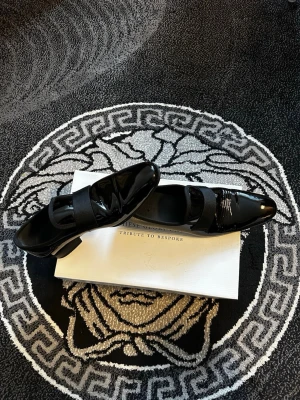 Svarta lackloafers med band - Säljer ett par stilrena svarta loafers i lackat läder med bred resår över vristen. Skorna har en elegant, spetsig tå och låg klack. Perfekta för dig som vill ha en dressad look med lite extra glans. Klassisk design som passar till många outfits.