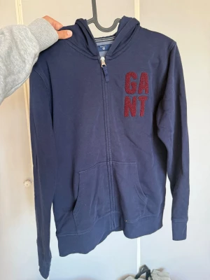 Marinblå hoodie från GANT - Snygg marinblå hoodie från GANT med dragkedja framtill och huva. På bröstet finns ett broderat GANT-märke i vinrött. Hoodien har känguruficka och är gjord i mjukt bomullsmaterial, perfekt för en avslappnad stil.