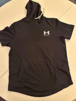 Svart Under Armour hoodie kort ärm - Svart hoodie från Under Armour med korta ärmar och vit logga på bröstet. Tröjan har huva med vit dragsko och en stor magficka framtill. Perfekt för en sportig och avslappnad stil. Materialet känns mjukt och bekvämt.