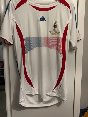 Frankrike matchtröja Henry 12 Adidas - Säljer en vit matchtröja från Adidas för Frankrikes landslag, med röda och blå detaljer samt tryck på bröstet och ryggen. På ryggen står det 'Henry' och nummer 12 i blått. Tröjan har korta ärmar och är tillverkad i ett lätt funktionsmaterial.