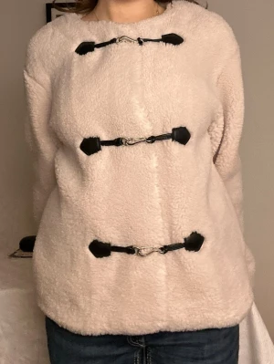 Beige teddyjacka med spännen🧸 - Mysig beige teddyjacka med tre svarta dekorativa spännen framtill. Jackan har rund halsringning och är tillverkad i fluffigt teddy-material som ger en trendig look. Perfekt för dig som vill ha en varm och stilren jacka med unika detaljer. Denna är superelegant och använd max 5 gånger!🧸