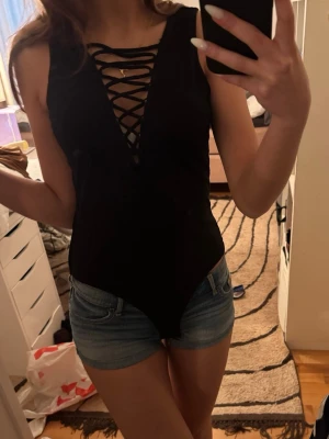 Svart body med snörning framtill - Säljer en svart body med djup v-ringning och snygg snörning framtill. Ärmlös modell i stretchigt material som sitter tight och bekvämt. Perfekt att styla med jeans eller kjol för en trendig look.