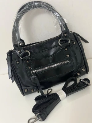 Svart handväska med nitar och dragkedja - Cool svart handväska i glansigt konstläder med silverfärgade nitar och detaljer. Väskan har både handtag och avtagbar axelrem, samt en dragkedjeficka framtill. Perfekt för dig som gillar en edgy och trendig look.