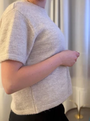 Beige stickad t-shirt i ullmix - Mysig beige stickad t-shirt med korta ärmar och rund hals. Tröjan har en rak passform och är tillverkad i en mjuk ullblandning som känns skön mot huden. Perfekt till alla outfits. 