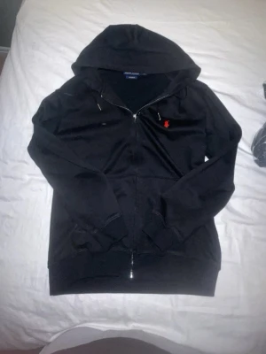 Svart hoodie från Polo Ralph Lauren - Svart hoodie med dragkedja från Polo Ralph Lauren. Klassisk design med huva och fickor framtill. Märkets ikoniska röda logga broderad på bröstet. Tillverkad i mjukt bomullsmaterial som känns skönt mot huden. Perfekt för en avslappnad och stilren look.
