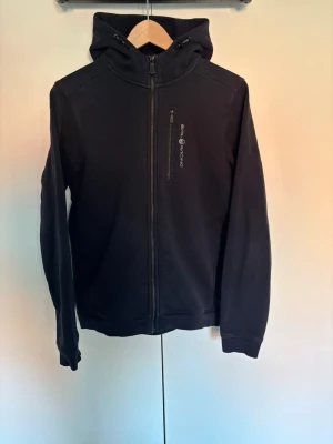 Svart hoodie från Sail Racing - Säljer en svart hoodie från Sail Racing med dragkedja framtill och en bröstficka med logga. Hoodien har en klassisk huva och diskret logotyp även på baksidan av huvan. Perfekt för dig som gillar stilren och sportig look. Hör av er vid frågor eller lägg ett prisförslag! 