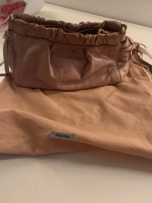 Rosa handväska från Miu Miu - Söt rosa handväska från Miu Miu i mjukt skinn med rynkade detaljer upptill och guldfärgade metalldetaljer. Väskan har dragkedja och dekorativa band på sidorna. Kommer med original dustbag och axel band 🎀 