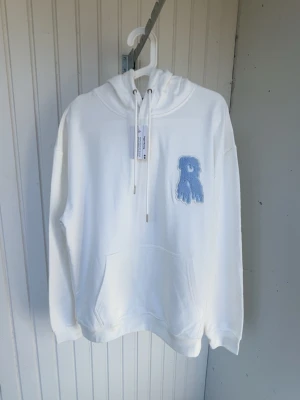 Vit hoodie från Risky med blå patch - Vit hoodie från Risky med stor huva och känguruficka. På bröstet sitter en blå patch i form av ett R med drip-effekt. Tröjan har långa ärmar och snörning vid halsen. Mjuk bomullskänsla och clean look, perfekt för dig som gillar streetwear. ( Helt ny, aldrig använt) väldigt sällsynt tröja 