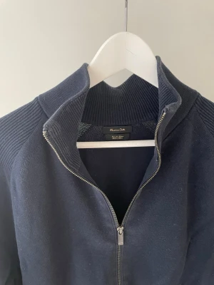 Mörkblå ziptröja från Massimo Dutti - Mörkblå ziptröja från Massimo Dutti med hög krage och hel dragkedja framtill. Tröjan har ribbade detaljer på axlar och krage samt långa ärmar. Perfekt för en stilren och avslappnad look.
