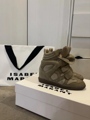Isabel Marant Bekett taupe sneakers - Säljer ett par Isabel Marant Bekett sneakers i taupe. De har en chunky siluett, dold kilklack och breda kardborreband. Skorna är gjorda i mjuk mocka och skinn, med gummisula. Perfekta för dig som gillar statement-sneakers med cool design.