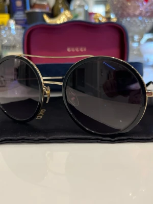 Runda svarta solglasögon från Gucci nyskick  - Snygga runda solglasögon från Gucci med svart båge och tunna guldfärgade detaljer. Glasen är mörka och bågarna har en elegant metallfinish. På skalmarna finns en liten guldfärgad bi-detalj som ger extra lyxig känsla. Perfekt accessoar för att höja din stil.