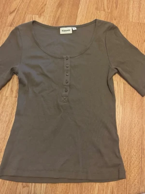 Brun ribbad topp från Kappahl - Snygg brun ribbad topp från Kappahl med rund halsringning och knappar framtill. Modellen har halvlånga ärmar och är tillverkad i mjuk bomull som sitter bekvämt mot kroppen. Perfekt att styla med jeans eller kjol.