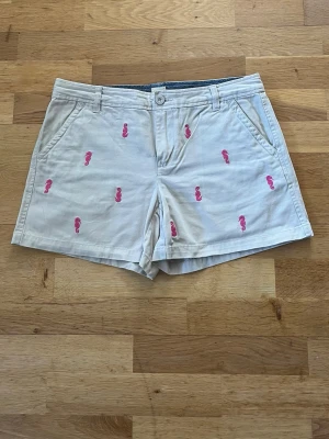 Beige shorts - Snygga beige shorts med rosa sjöhästar broderade över hela plagget. Klassisk passform med fickor fram och bak samt bälteshällor. Perfekta för sommaren och tillverkade i mjuk bomull för skön känsla.