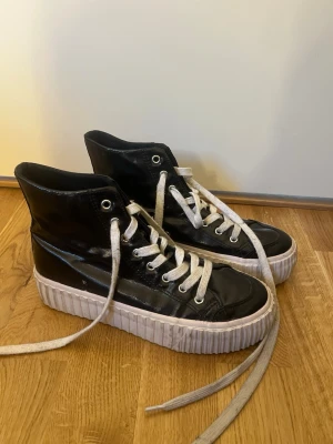 Svarta höga sneakers med platåsula - Säljer ett par svarta höga sneakers med vit platåsula och vita skosnören. Skorna har en glansig finish och metallfärgade öljetter. Snygg och edgy look som passar till streetwear och ger extra höjd. Klassisk snörning framtill och robust sula.