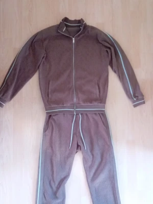 Mjukisdress - Säljer ett brunt loungewear-set med matchande ziptröja och byxor. Setet har ljusblå ränder längs ärmar och ben samt ribbade muddar och midja. Materialet ser ut att vara mjukt och bekvämt, perfekt för att chilla hemma. Dragkedja framtill på tröjan och dragsko i midjan på byxorna.