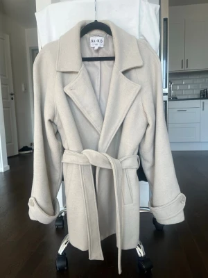 Beige kappa från NA-KD med skärp - Stilren beige kappa från NA-KD med bred krage, knytskärp i midjan och uppvikta ärmslut. Kappan har en klassisk rak passform och är fodrad för extra komfort. Perfekt för dig som vill ha en tidlös och trendig look.