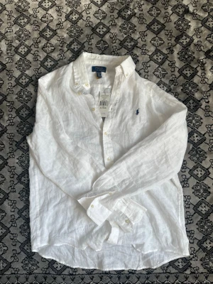 Ralph lauren skjorta - En vit Ralph Lauren skjorta i linne som är perfekt nu för sommaren som är runt hörnet. Den är väldigt luftig och passar perfekt till båda stranden och en lungt kompis häng. Storlek 18-20 skulle säga att detta motsvarar S-M i normal storlek. Helt ny aldrig använd. 100% riktig med qr kod