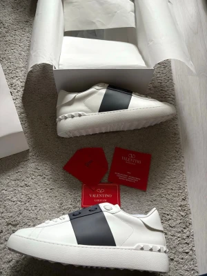 Valentino Garavani vita sneakers - Snygga vita sneakers från Valentino Garavani med bred mörkblå rem över ovandelen. Skorna har en texturerad sula och nitar vid hälen för en cool detalj. Tillverkade i läder med klassisk låg profil och stilren design. Storlek 42
