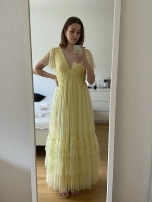 Ljusgul långklänning med volanger - Superfin ljusgul långklänning med v-ringning och korta ärmar i tunn, luftig tyll. Klänningen har flera lager volanger nedtill och markerad midja som ger en romantisk och flowy look. Perfekt för sommarens alla tillfällen!