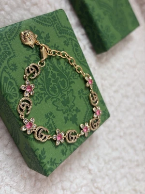 Guldigt armband med rosa stenar - Snyggt armband i guldfärgad metall med dekorativa länkar och detaljer av rosa och vita stenar i blommönster. Armbandet har en kedja med justerbar längd och ett unikt lås med lejonmotiv. Perfekt accessoar för att lyfta din outfit, (inspirerat).