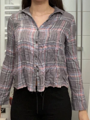 Grå-rosa rutig skjorta med knappar - Säljer en grå-rosa rutig skjorta med långa ärmar och knappar framtill. Skjortan har en avslappnad passform och är tillverkad i ett lätt bomullstyg. Perfekt för dig som gillar en chill och trendig stil.