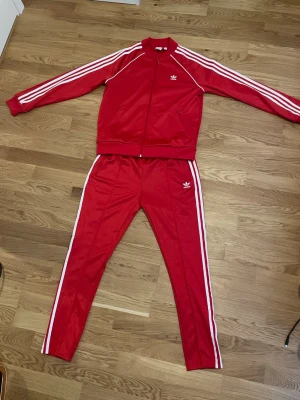 Röd SST Adidas träningsdress - Säljer en klassisk röd Adidas träningsdress med vita ränder längs ärmar och ben. Setet består av zip-jacka och matchande byxor, båda med Adidas logga på bröst och höft. Tillverkad i glansigt syntetmaterial, perfekt för sport eller chill.