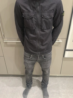 Svart vindjacka från Stone Island - Säljer en svart vindjacka från Stone Island med ikonisk patch på ärmen. Jackan har två bröstfickor med knappar, dragkedja framtill och en clean, stilren look. Perfekt för dig som gillar streetwear och vill ha något snyggt och funktionellt. Passar till 165 cm.