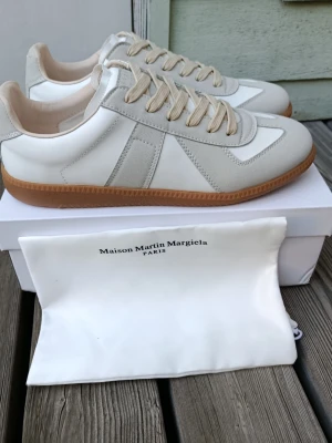 Maison Margiela vita sneakers - Säljer ett par stilrena vita sneakers från Maison Martin Margiela med beige mockadetaljer och gummisula. Skorna har klassisk låg silhuett, vita och beige partier i läder och mocka samt matchande beige skosnören. Perfekta för en clean och trendig look.