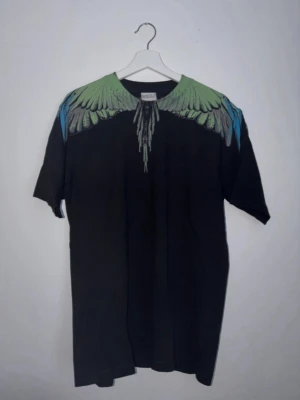 Marcelo Burlon svart t-shirt med vingar - Säljer en svart t-shirt från Marcelo Burlon med ikoniskt vingtryck i grönt, blått och grått över axlar och bröst. T-shirten har normal passform och är tillverkad i mjuk bomull. Perfekt för dig som vill sticka ut med en unik design.