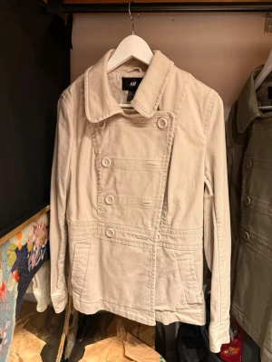 Beige dubbelknäppt rock från H&M - Stilren beige rock från H&M med dubbelknäppning och bred krage. Tillverkad i ett mjukt bomullstyg och har två fickor framtill samt dekorativa knappar bak. Perfekt för dig som gillar en klassisk men avslappnad look.
