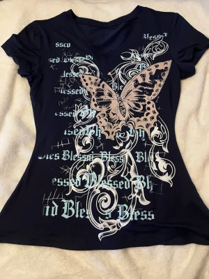Mörk blå t-shirt med fjärilstryck - Cool mörk blå t-shirt med v-ringning och ett stort beige fjärilstryck framtill. T-shirten har även vita och ljusblå grafiska detaljer och texten 'Blessed' i olika stilar.