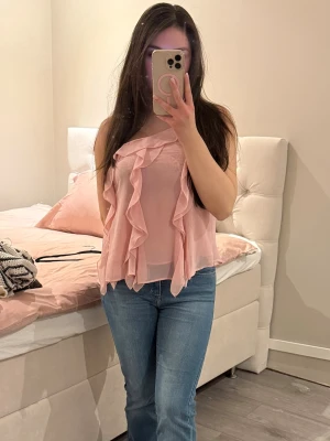 Ljusrosa volangtop från Zara - Säljer en ljusrosa, asymmetrisk topp från Zara med tunna axelband och volangdetaljer framtill. Toppen är gjord i ett lätt, transparent tyg som ger ett luftigt intryck och har en trendig, flowy passform. Blusen är endast använd få gånger 🌸