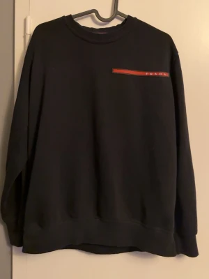 Svart sweatshirt från Prada - Svart sweatshirt från Prada med rund halsringning och diskret röd logga på bröstet. Tröjan har lång ärm och en klassisk, avslappnad passform. Perfekt för dig som gillar stilrena och exklusiva plagg.