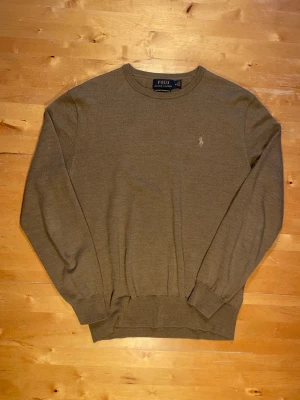 Olivgrön tröja från Polo Ralph Lauren - Snygg olivgrön tröja från Polo Ralph Lauren med rund halsringning och diskret broderad logga på bröstet. Tillverkad i mjuk och tunn ullmix, perfekt för lager på lager. Klassisk passform med ribbade muddar vid ärmslut och nederkant.