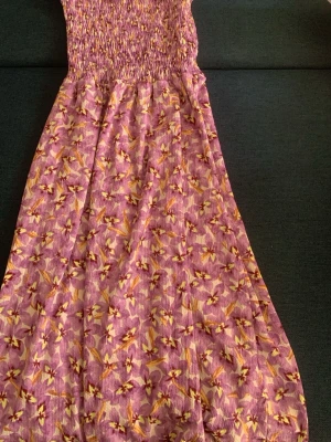 Lång blommig klänning i lila toner - Säljer en lång klänning i lila med blommigt mönster i gult och vitt. Klänningen har smock upptill och ett flowy fall. Perfekt för varma dagar och har en somrig vibe. Materialet känns lätt och luftigt.