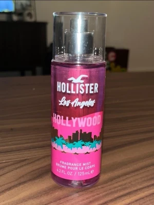 Hollister Hollywood Body Mist (Oanvänd) - En snygg body mist från Hollister med namnet Hollywood. Flaskan är cylinderformad och genomskinlig med rosa och lila toner, dekorerad med palmer och stadssilhuett. Innehåller 125 ml och har en praktisk sprayfunktion. Perfekt för dig som gillar fräscha och ungdomliga dofter.