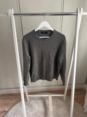 Grå stickad tröja från Polo Ralph Lauren - Klassisk grå stickad tröja från Polo Ralph Lauren med rund halsringning och ribbade muddar. Tröjan har en liten grön broderad logga på bröstet och är tillverkad i mjukt material, 100% bomull. Gott skick, passar till det mest.