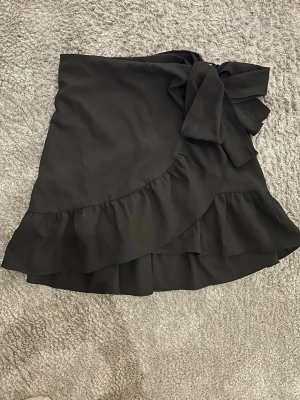 Svart omlottkjol med volang!! - Söt svart kort omlottkjol med volangkant och stor knytning i midjan! Inga skador, som ny!!💗💗💗