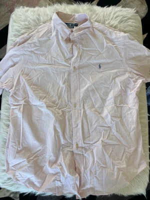 Rosa- och vitrandig skjorta Ralph Lauren - Kortärmad skjorta från Ralph Lauren i ljusrosa och vitt med tunna ränder. Klassisk krage, knappar framtill och den ikoniska blå loggan broderad på bröstet. Tillverkad i bomull för en sopren och luftig känsla.