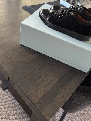 Svarta sneakers med lackdetaljer - Snygga svarta lanvins sneakers med glansiga lackdetaljer på tån och klassisk snörning. Ovandelen verkar vara i mocka eller liknande material och sulan är svart. Perfekta för dig som vill ha en stilren och trendig look. Storlek 40 men passar även 41