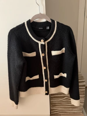 Svart kofta med vita detaljer Vero Moda - Stilren svart kofta från Vero Moda med vita ribbade kanter och dekorativa fickor. Rund halsringning, guldknappar framtill och mjukt stickat material. 