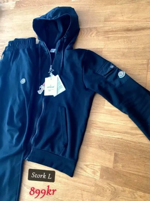 Svart Moncler loungewear set - Snyggt svart loungewear set från Moncler med dragkedja, huva och fickor. Setet har Moncler-logga på både byxa och ärm samt en liten ficka med knapp på ärmen. Tillverkat i mjukt material som passar perfekt för chill dagar hemma.