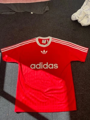  röda Adidas Originals-tshirt med vita detaljer - Klassisk röd t-shirt från Adidas med vita ränder på axlarna och stort Adidas-tryck framtill. T-shirten har rund halsringning med vit kant och är kortärmad. Perfekt för en sportig och avslappnad stil.