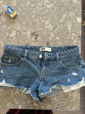 Blå jeansshorts från JDY - Säljer ett par blå jeansshorts från JDY i modellen Small. Shortsen har råa, fransiga kanter och slitna detaljer framtill. Klassisk femficksmodell med knapp och dragkedja. Perfekta för varma dagar och en avslappnad stil. Helt slutsålda 