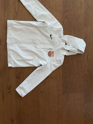 Vit Nike FC Barcelona vindjacka - Snygg vit vindjacka från Nike med FC Barcelona-logga på bröstet. Jackan har huva med dragsko, hel dragkedja framtill och två sidofickor. Perfekt för dig som vill ha sportig stil och visa ditt lagstöd.